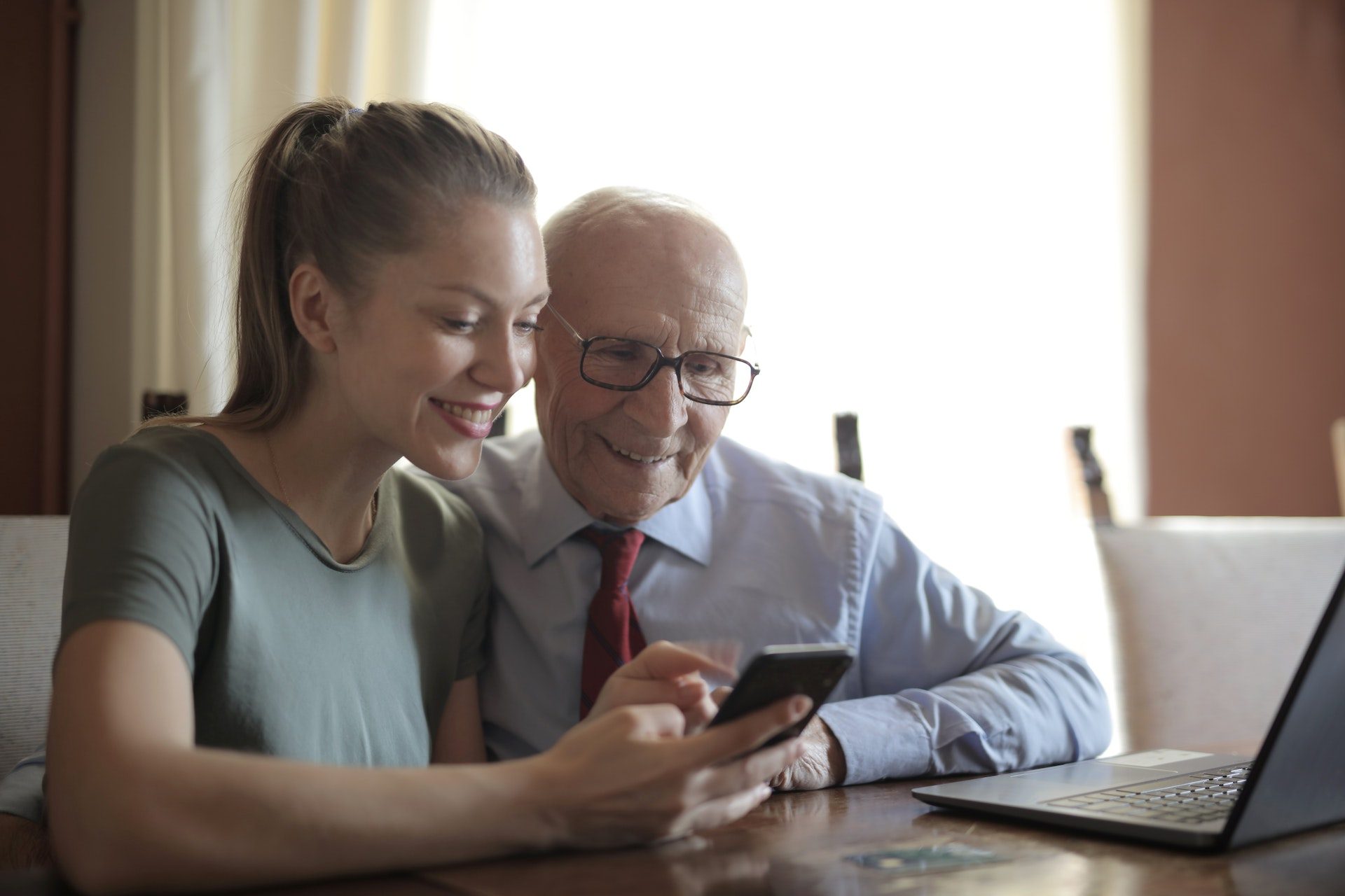 Smiling young woman in casual clothes showing smartphone to interested senior grandfather in formal shirt and eyeglasses while sitting at table near laptop